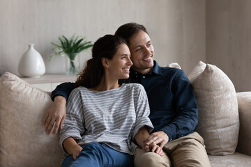 Happy married couple relaxing on couch at home, looking away, talking, thinking over future family, planning house buying, mortgage, apartment rent. Smiling dreamy husband embracing wife