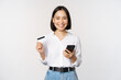Online shopping concept. Image of young asian modern woman holding credit card and smartphone, buying with smartphone app, paying contactless, standing over white background