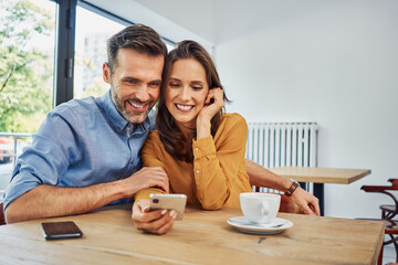 Wall Mural - Happy couple looking at smartphone sitting together in cafe browsing together social media