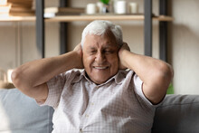Happy Positive Elderly Senior Man With Closed Eyes Covering Ears With Hands With Toothy Smile, Sitting On Sofa, Playing Hide And Seek With Grandchildren. Front View, Head Shot Portrait