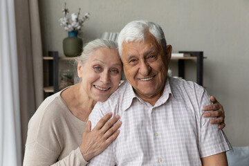 Wall Mural - Happy elderly 70s 80s couple posing in home interior head shot portrait. Old grey haired husband and wife hugging with love, tenderness, care, looking at camera together with toothy smiles, laughing