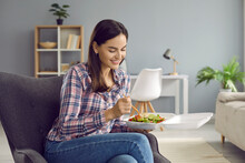 Pretty woman having takeout lunch at home. Happy young girl sitting in armchair in living room, holding plastic takeaway food container and eating veggie salad with plastic fork. Meal delivery concept