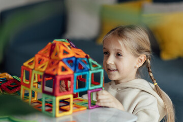Candid lifestyle portrait of Cute caucasian Kid eight years old playing with magnetic constructor toy and building house from blocks at home. Little child girl plays with colorful blocks and