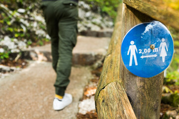 Blue round sign on a wooden fence by a park trail informing people to keep 2 meter 6 feet physical social distance from each other,prevent & protect Coronavirus COVID-19 virus disease infection spread