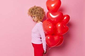 Wall Mural - Offended young woman with curly hair stands sideways at camera holds bunch of inflated balloons behind back feels stressed and lonely isolated over pink background. Sad Valentines Day concept