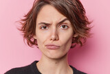 Fototapeta  - Headshot of serious beautiful young woman looks skeptical raises eyebrows purses lips considers something tries to make decision isolated over pink background. Human face expressions concept