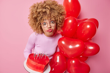 Wall Mural - Upset curly haired young woman feels lonely on Valentines Day holds bunch of heart shaped balloons and delicious cake wears transparent glasses has spoiled makeup poses against pink background