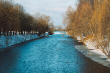 Blue River With Snow Ducks And Trees With A Blue Bridge And Dark Sky In The Background