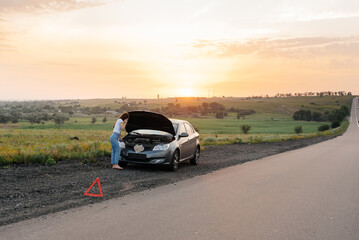 Wall Mural - A young girl stands near a broken car in the middle of the highway during sunset and tries to call for help on the phone and start the car. Waiting for help. Car service. Car breakdown on the road.