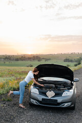 Wall Mural - A young girl stand near a broken car in the middle of the highway during sunset and tries to repair it. Troubleshooting the problem. Car service. Car breakdown on the road.