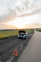 Wall Mural - A young girl stands near a broken car in the middle of the highway during sunset and tries to repair it. Troubleshooting the problem. Waiting for help. Car service. Car breakdown on road..