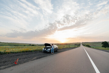 Wall Mural - A young girl stands near a broken-down car in the middle of the highway during sunset and tries to call for help on the phone. Waiting for help. Car service. Car breakdown on road.