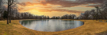 A Panoramic Shot Of A Still Green Lake In The Park Surrounded By Yellow Winter Grass, Bare Winter And Lush Green Trees And Smooth Footpath And A Yellow Sky At Sunset At MLK Riverside Park In Memphis
