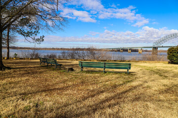 two green wooden benches on yellow winter grass along the Mississippi river near the Memphis–Arkansas Memorial Bridge with bare winter trees blue sky and powerful clouds at Mud Island Park in Memphis 