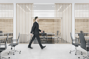 Poster - Side view of businessman walking in modern wooden and concrete office interior with furniture and blinds. Design and architecture concept.