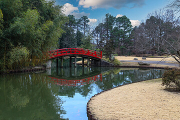 Wall Mural - a shot of an arched red and black bridge over a lake surrounded lush green trees reflecting off the water with blue sky and powerful clouds at Memphis Botanic Garden in Memphis Tennessee USA