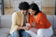 Smiling couple using mobile phone shopping online, ordering food sitting at home. Happy relaxed African American woman and Indian man holding smartphone communication, looking at digital screen