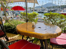 Summer Cafe In Yacht Club. Restaurant Furniture. Table Made Of Lacquered Solid Saw Cut Tree Trunk. Stylish Interior. Wicker Chairs, Bright Red Cushions And Parasols. Moored Luxury Expensive Yachts. 