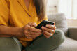 © shurkin_son - Crop shot of elderly woman's hands holding black mobile phone against background of her yellow blouse and green jeans, sitting on couch, spending leisure time in social networks, texting message