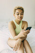 © shurkin_son - Charming African young curly blonde female in light green bodysuit wearing nose and ears jewerly using smartphone for chatting with her beloved, looking romantic and calm sitting on bed in morning