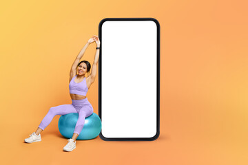 Wall Mural - Fitness app. Athletic black lady training on fitness ball near big cellphone with blank screen, mockup