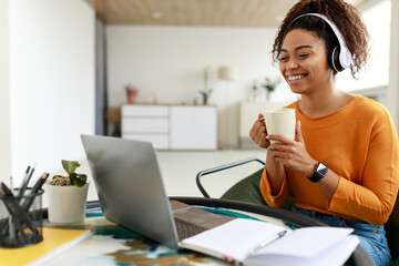 Canvas Print - Smiling black lady watching webinar on computer, drinking hot coffee