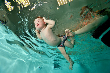 Mom teaches swimming and helps a child with Down syndrome swim underwater in a children's pool with blue water. Children's disability. Portrait. Shooting from the bottom. Horizontal orientation.