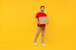 Full body smiling happy delivery guy employee man in red cap T-shirt uniform workwear work as dealer courier hold cardboard box isolated on plain yellow background studio portrait. Service concept