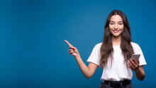 Attractive Caucasian Or Arab Brunette Girl In A White T-shirt Using A Smartphone And Pointing Her Finger At A Copy Space For Advertising Isolated On A Blue Studio Background.
