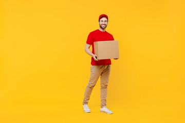 Wall Mural - Full body smiling happy delivery guy employee man in red cap T-shirt uniform workwear work as dealer courier hold cardboard box isolated on plain yellow background studio portrait. Service concept