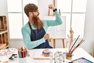 Poster - Redhead man with long beard painting clay bowl at art studio showing arms muscles smiling proud. fitness concept.