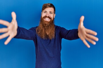 Poster - Redhead man with long beard wearing casual blue sweater over blue background looking at the camera smiling with open arms for hug. cheerful expression embracing happiness.