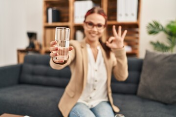 Sticker - Young caucasian woman offering a glass of water doing ok sign with fingers, smiling friendly gesturing excellent symbol