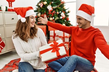 Sticker - Young hispanic couple wearing christmas hat opening gift at home.