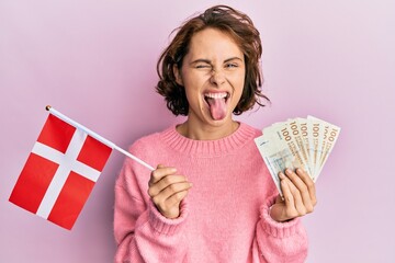 Sticker - Young brunette woman holding norway flag and krone banknotes sticking tongue out happy with funny expression.