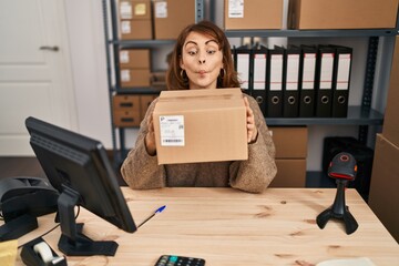 Wall Mural - Young beautiful woman working at small business ecommerce holding box making fish face with mouth and squinting eyes, crazy and comical.