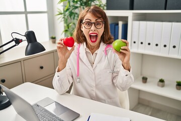 Sticker - Young doctor woman holding heart and green apple celebrating crazy and amazed for success with open eyes screaming excited.