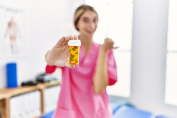 Sticker - Young physiotherapist woman working at pain recovery clinic holding pills pointing thumb up to the side smiling happy with open mouth