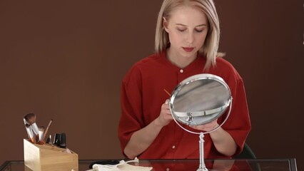 Poster - Beautiful blond hair woman in red shirt applying makeup in opposite of mirror