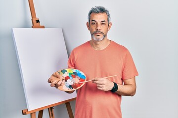 Poster - Handsome middle age man with grey hair standing drawing with palette by painter easel stand relaxed with serious expression on face. simple and natural looking at the camera.