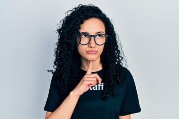 Young hispanic woman with curly hair wearing staff t shirt thinking concentrated about doubt with finger on chin and looking up wondering