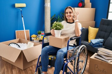 Poster - Young woman sitting on wheelchair moving to a new home smiling positive doing ok sign with hand and fingers. successful expression.