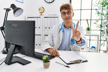 Canvas Print - Young caucasian doctor man working at the clinic showing and pointing up with fingers number two while smiling confident and happy.