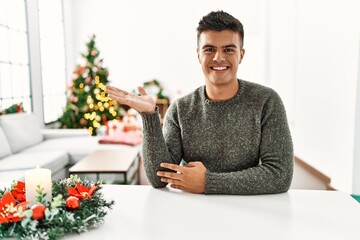 Canvas Print - Young hispanic man sitting on the table by christmas tree smiling cheerful presenting and pointing with palm of hand looking at the camera.