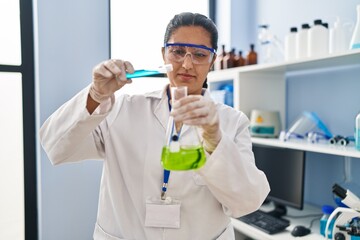 Wall Mural - Young hispanic woman wearing scientist uniform pouring liquid on test tube at laboratory