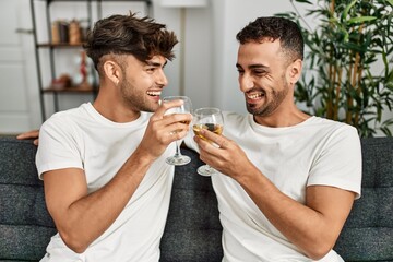 Wall Mural - Two hispanic men couple toasting with glass of wine sitting on sofa at home