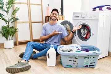 Wall Mural - Young hispanic man putting dirty laundry into washing machine inviting to enter smiling natural with open hand