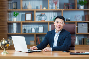 Wall Mural - Successful asian businessman looking at camera and smiling, man working in classic office at desk, with laptop, looking at camera portrait