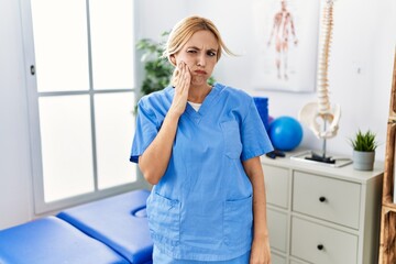 Poster - Beautiful blonde physiotherapist woman working at pain recovery clinic touching mouth with hand with painful expression because of toothache or dental illness on teeth. dentist