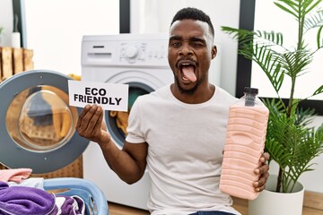 Wall Mural - Young african man doing laundry holding eco friendly paper sticking tongue out happy with funny expression.